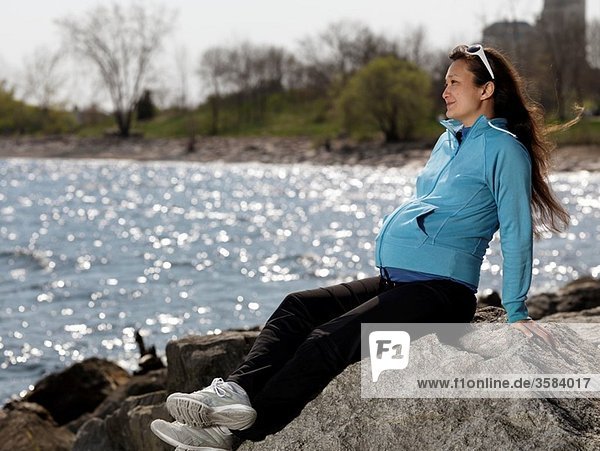 Pregnant young woman sitting at a rocky shore near water