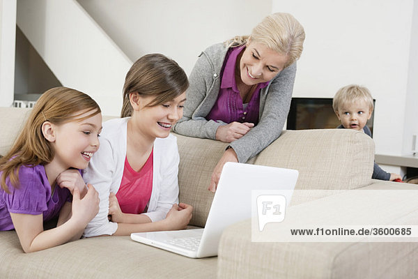 Zwei Mädchen  die einen Laptop auf einer Couch benutzen  während ihre Großmutter neben ihnen steht.