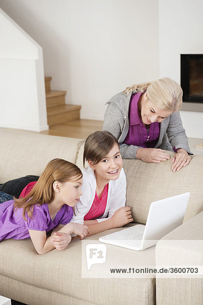 Zwei Mädchen  die einen Laptop auf einer Couch benutzen  während ihre Großmutter neben ihnen steht.