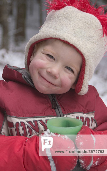 Junge 4 Jahre alt lacht im Schnee und trinkt einen warmen Tee aus einem Becher