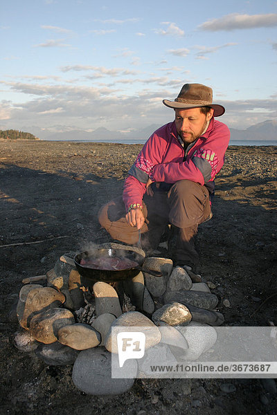 Ein Mann bereitet am Strand mit einer Gußeisenpfanne über dem offenen Feuer ein Steak zu Alaska USA