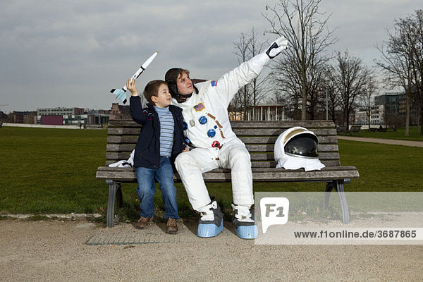 Ein Junge mit einer Modellrakete  der mit einem Astronauten auf einer Parkbank sitzt.
