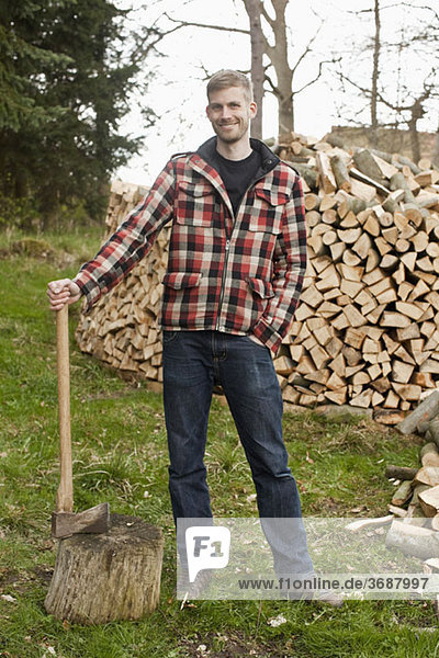 Ein Mann  der eine Axt hält und vor einem Holzhaufen steht.