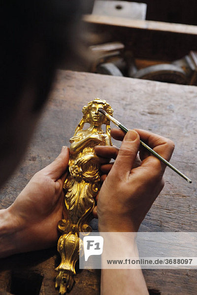 Detail einer Person  die eine vergoldete Figur und einen Pinsel hält.