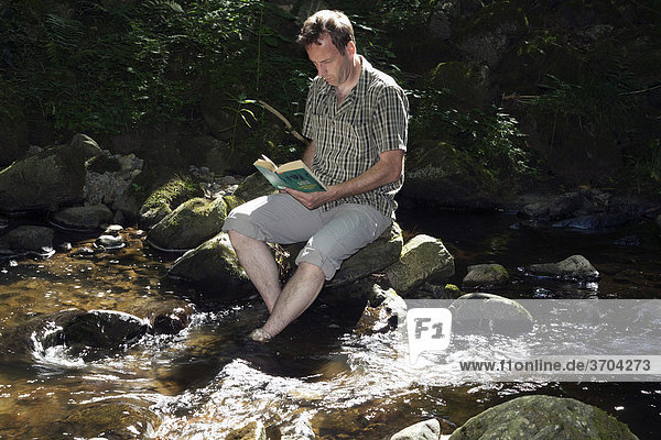Male  mid 40s  sitting in a stream and reading a book  Hoellbach  Black Forest  Germany  Europe