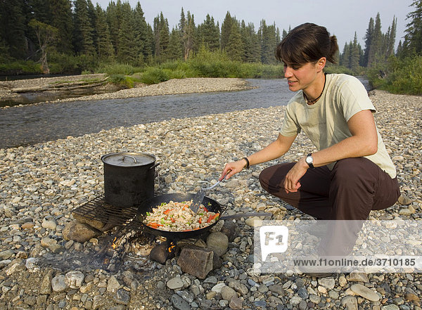 Young woman cooking  frying chicken stir fry on a camp fire  gravel bar  upper Liard River  Yukon Territory  Canada