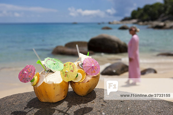 Zwei dekorierte Kokosnüsse mit einem Getränk gefüllt stehen auf einem Granitfelsen  hinten steht eine Frau in einer rosa-farbenen Tunika  Insel Mahe  Seychellen  Indischer Ozean  Afrika