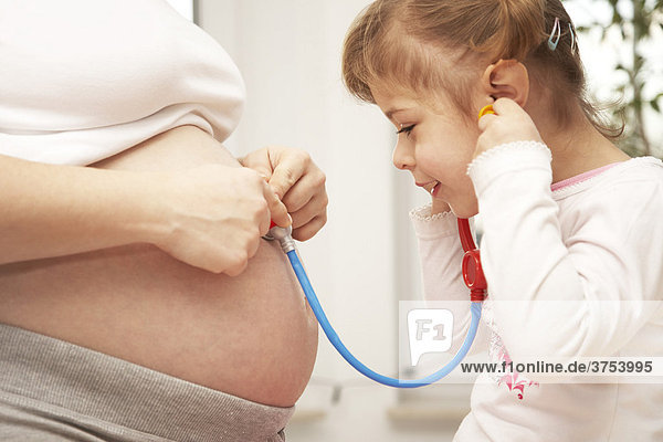 Vierjährige Tochter hört mit Stethoskop den Schwangerschaftbauch der Mutter ab