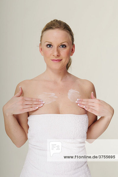 Blonde young woman wrapped in a white towel applying body lotion