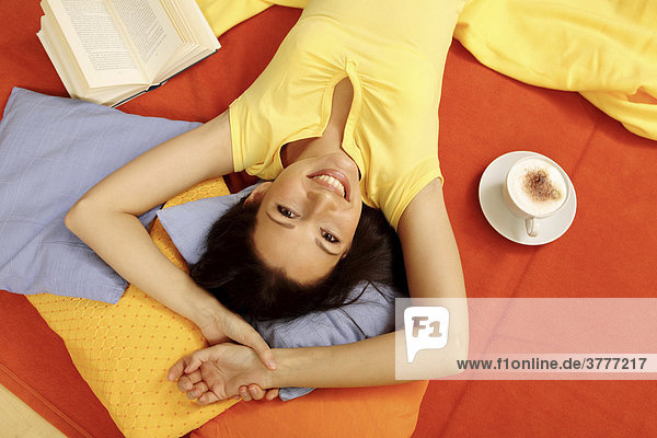Junge Frau räkelt sich mit Milchkaffee und Buch auf bunten Kissen