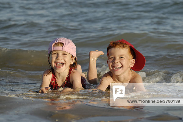 Zwei Kinder liegen am Strand im Wasser und freuen sich