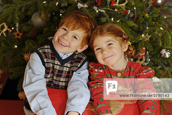 Zwei Kinder sitzen mit einem Geschenk unter dem Weihnachtbaum