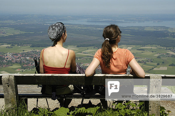 Rücken von zwei jungen Frauen sitzen auf einer Bank mit Blick zum Chiemsee am Berg Hochgern Chiemgau Bayern