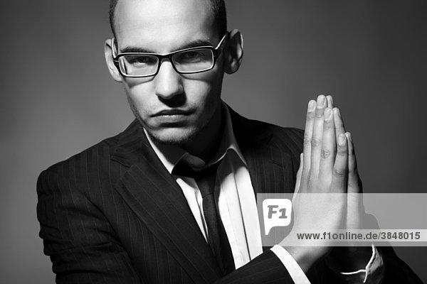 Portrait eines jungen Mannes mit Brille  Anzug  Hemd und Krawatte die Hände gefaltet neben sich haltend