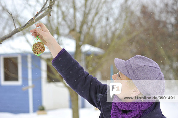 Frau hängt Beutel mit Erdnüssen als Winterfütterung für Vögel an einen Baum