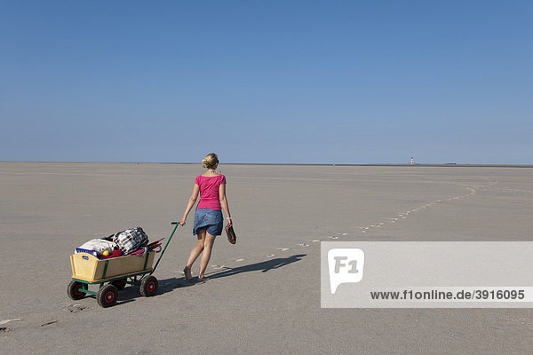 Junge Frau  20-25 Jahre  zieht ihren Bollerwagen mit Strandgepäck über den Sandstrand von St Peter Ording  Nordsee  Nordfriesland  Schleswig-Holstein  Norddeutschland  Deutschland  Europa