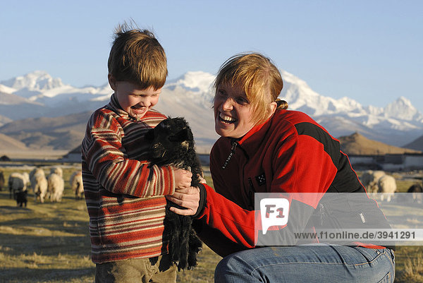 Kleines Mädchen  Kind  hält freudestahlend ein kleines schwarzes Schaf  Lamm  zusammen mit lachender Frau vor verschneitem Gebirgszug des Cho Oyo  8112 m  in der Hochebene von Tingri  Tibet  China  Asien