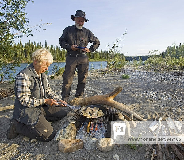 Woman cooking  frying fish fillets in a pan on a camp fire  man with plate waiting behind  upper Liard River  Yukon Territory  Canada
