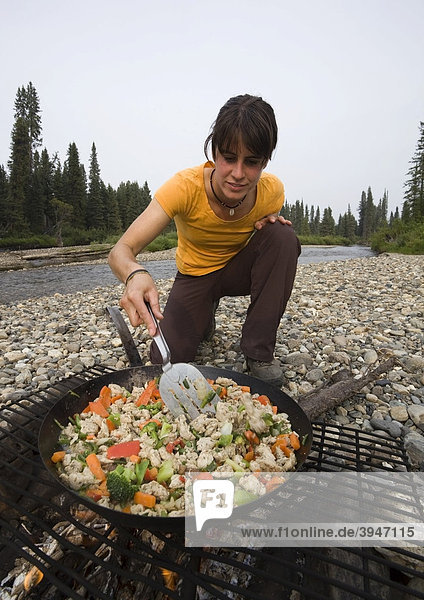 Young woman cooking  frying chicken stir fry on a camp fire  pan  spatula  gravel bar  upper Liard River  Yukon Territory  Canada