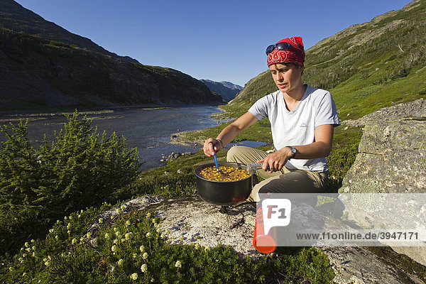 Junge Frau bereitet eine Mahlzeit zu  Kochen im Freien  Reisekocher  Happy Camp  historischer Chilkoot Pass  Chilkoot Trail Wanderweg  dahinter der Long Lake See  Yukon Territory  British Columbia  BC  Kanada