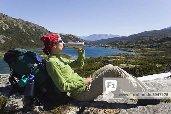 Junge Wanderin  Rucksacktourist  auf einem Felsen sitzend  trinkt  ruht aus und genießt das Panorama  Deep Lake See  historischer Chilkoot Pass  Chilkoot Trail Wanderweg  Yukon Territory  British Columbia  BC  Kanada