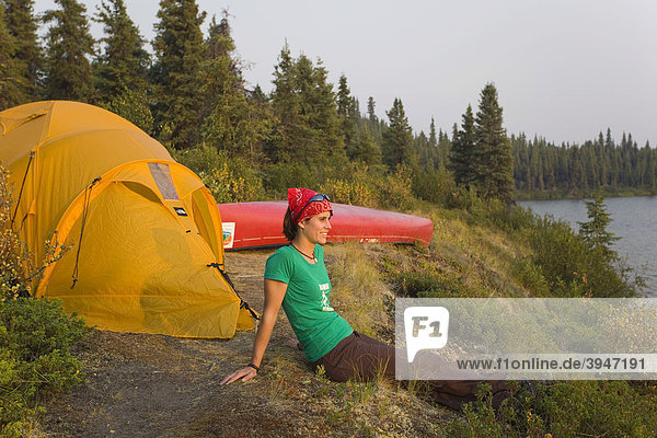 Young woman sitting  enjoying view  relaxing  tent and canoe behind  Caribou Lakes  upper Liard River  Yukon Territory  Canada