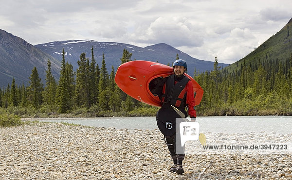 White water kayaking  man carrying a play boat  kayak and paddle  dry suit  helmet  mountains  coastal range behind  Wheaton River  Yukon Territory  Canada
