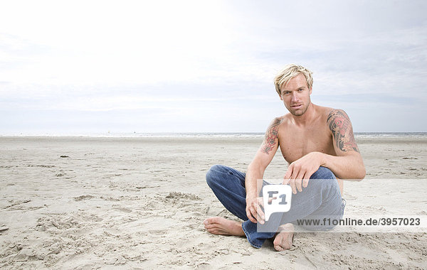 Junger Mann sitzt mit freiem Oberkörper am Strand