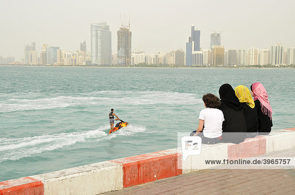 Verschleierte Frauen beobachten Wasserscooter-Fahrer vor der Skyline von Abu Dhabi  Vereinigte Arabische Emirate  Arabien  Naher Osten  Orient