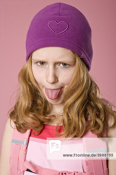 Rothaariges Mädchen mit violetter Mütze und Schultasche vor Rosa  streckt die Zunge heraus