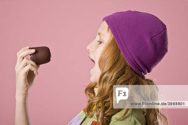 Rothaariges Mädchen mit violetter Mütze vor Rosa beißt in einen Schokokuss
