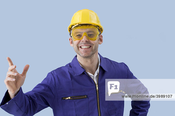 Handwerker im Blaumann mit Schutzbrille und gelbem Schutzhelm gestikuliert