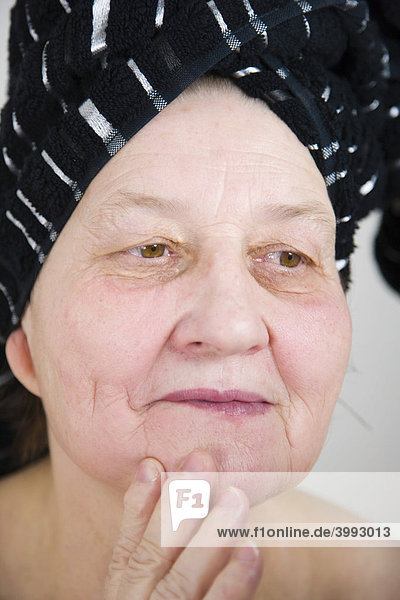 67 Jahre alte Frau mit einem Handtuch auf dem Kopf kontrolliert ihre Gesichtshaut