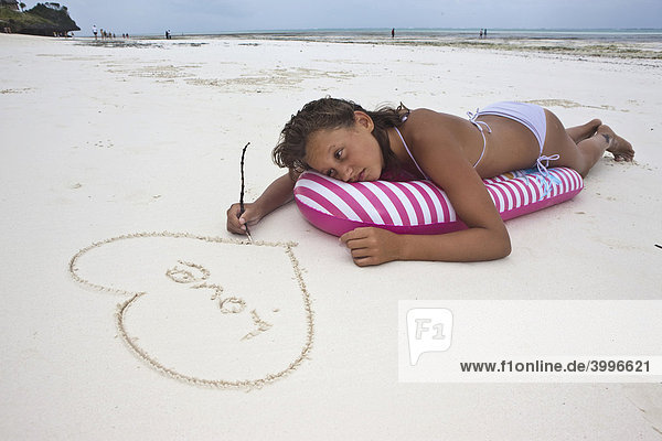 Ein Mädchen  ca 12 Jahre  mit Liebeskummer liegt im Urlaub auf einer Luftmatratze und malt ein Herz mit dem Wort Love in den Sand