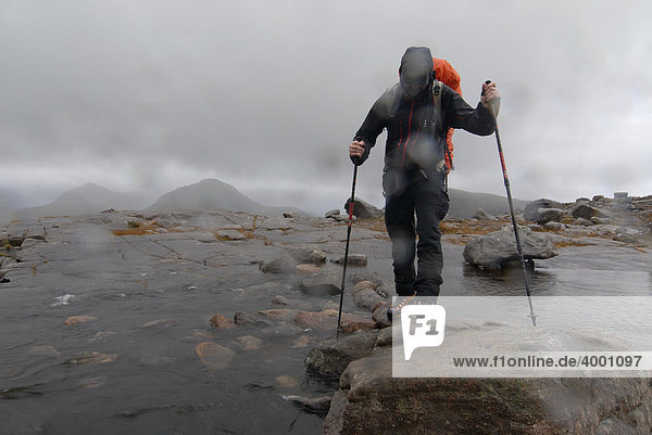 Wanderer mit Teleskopstöcken und Regenanzug beim Überqueren eines Flusses im schottischen Regen  schottische Highlands  Liathach  Torridon Schottland  Großbritannien  Europa