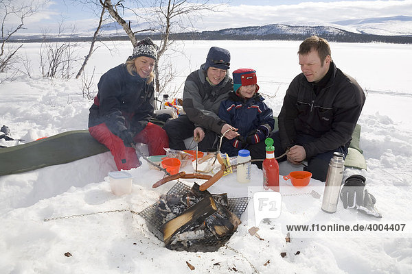 Grillparty in Lappland  ein Mann  ein vierjähriges Kind und zwei Frauen sitzen am Ufer des zugefrorenen Holmajärvi-Sees und halten Würste über das Feuer in Kiruna  Lappland  Nord-Schweden  Schweden