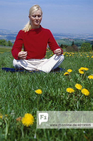 Blonde Frau mit rotem Pulli sitzt in der Wiese und meditiert