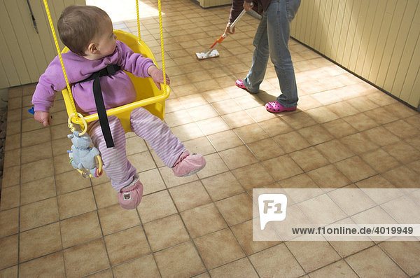 Baby girl in a swing looking over her shoulder at her mother mopping a tiled floor