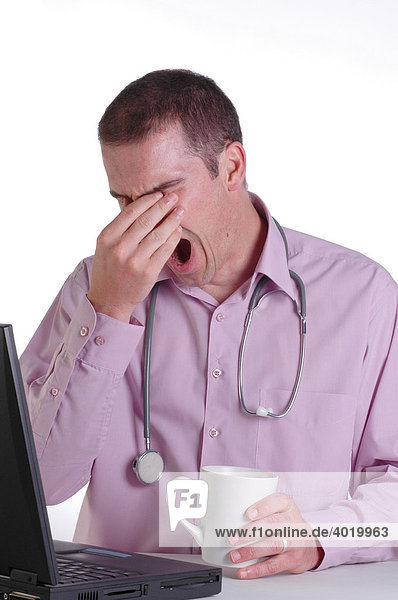 A young male doctor at his desk rubbing his eyes and yawning with fatigue from working long hours
