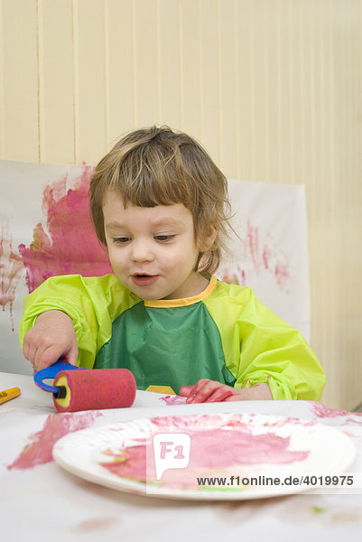 A two year old girl in a plastic smock painting in a playroom with a brush and roller