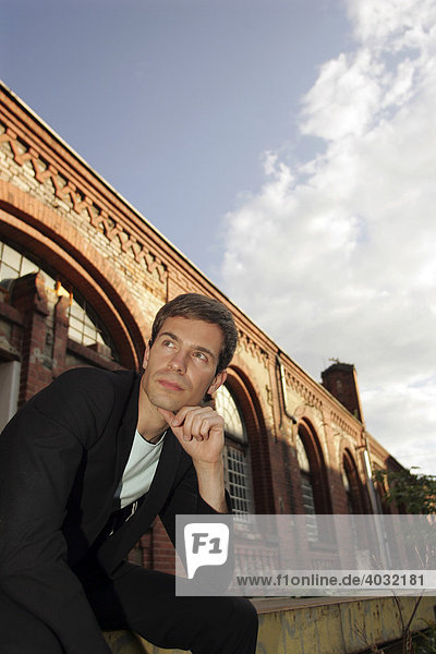 Portrait of a young man wearing a suit  sitting in front of an industrial building made of red bricks looking into the distance  thinking