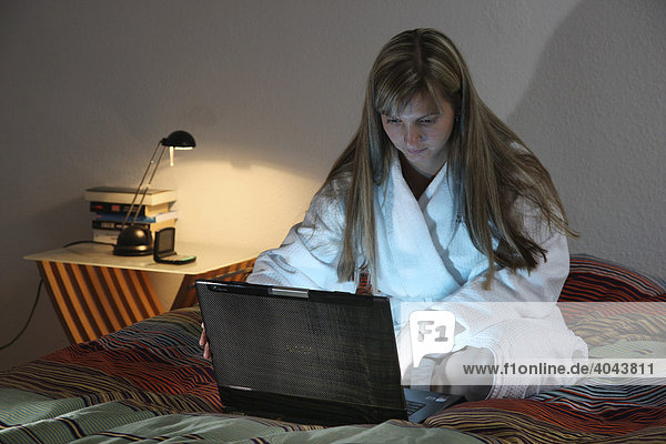 Junge Frau  25-30 Jahre  surft mit Laptop Computer im Internet im Bademantel auf dem Bett
