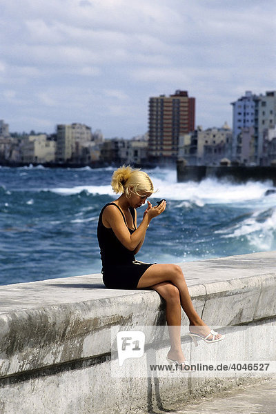Young blonde Cuban woman sitting on a wall at the sea  looking into a mirror  checking her makeup  Malecon  Havana  Cuba  Caribbean
