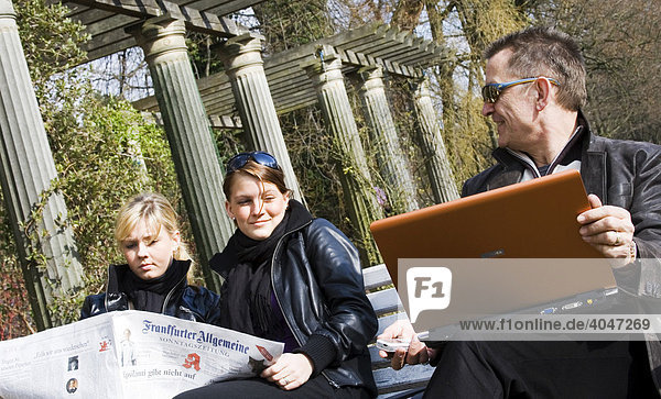 Mann sitzt mit Laptop neben zwei Teenies  Teenagern auf einer Bank im Park  Berlin  Deutschland  Europa