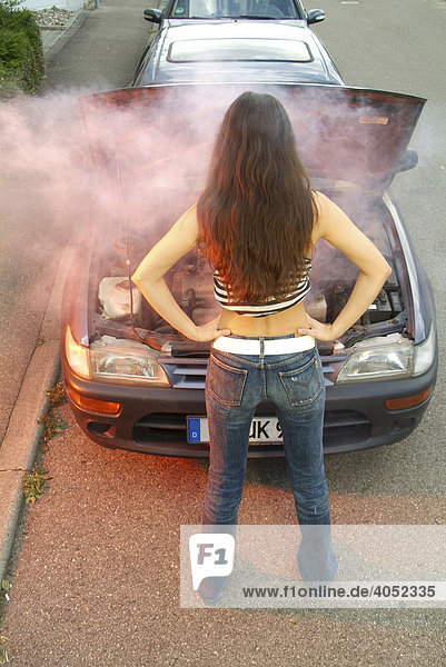 Junge Frau steht vor ihrem qualmenden Auto und schaut machtlos in den geöffneten Motorraum