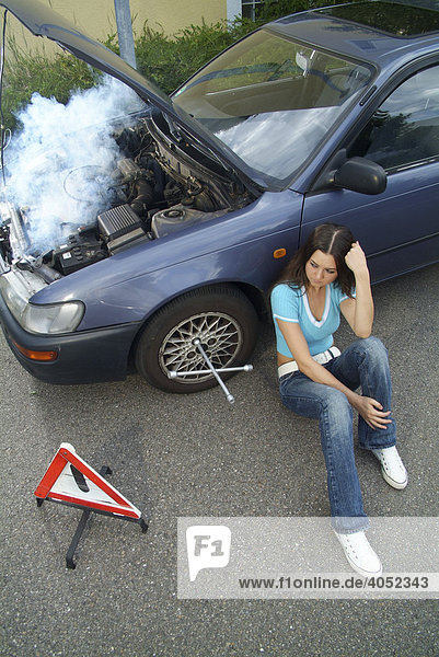 Frustrierte junge Frau mit Warndreieck sitzt an der Seite ihres defekten und qualmenden Autos und wartet auf Hilfe