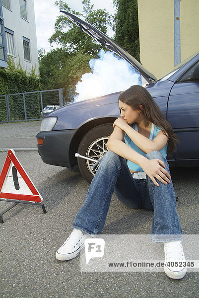 Frustrierte junge Frau mit Warndreieck sitzt an der Seite ihres defekten und qualmenden Autos und wartet auf Hilfe