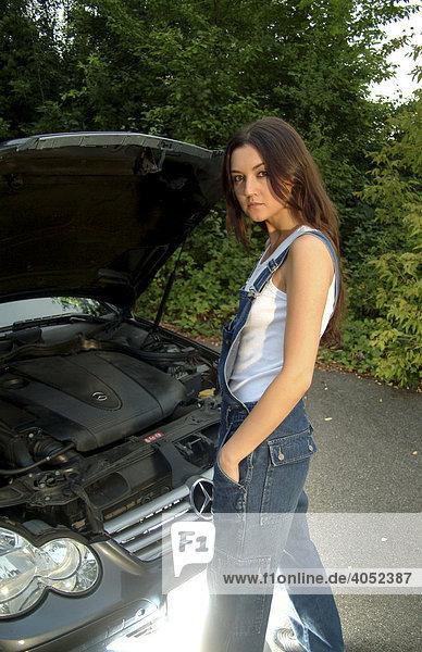 Junge Frau in Arbeitskleidung und mit geöffneter Motorhaube bei der Reparatur ihres Autos