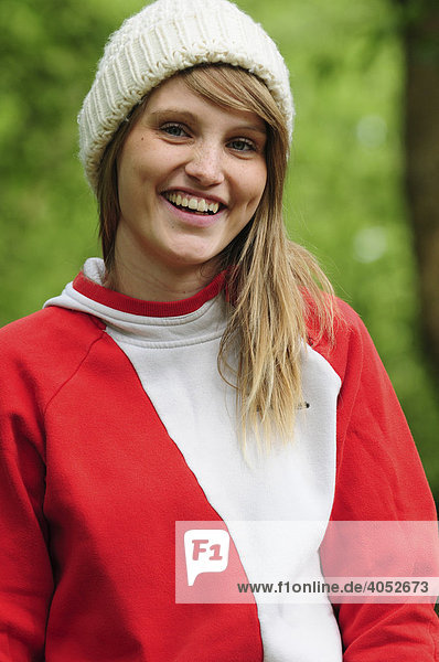 Portrait junge  schöne  sportliche  blonde Frau im Sommer im Park  lächelt  trägt Mütze und roten Kapuzenpullover