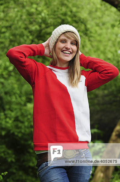 Portrait junge  schöne  sportliche  blonde Frau im Sommer im Park  lacht  trägt Mütze und roten Kapuzenpullover